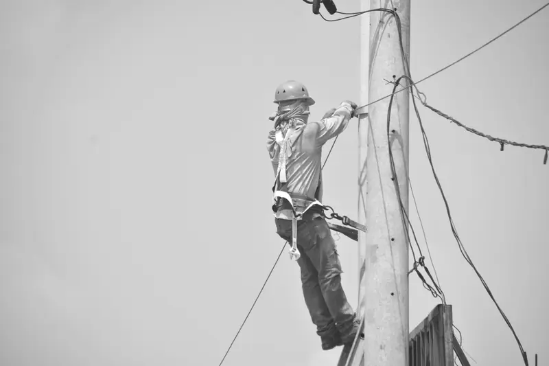 Liniero con casco y arnés de posicionamiento asegurado a un poste de electricidad (blanco y negro)