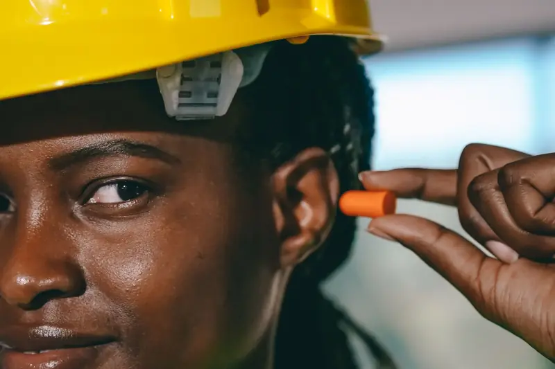 Trabajadora con casco amarillo insertándose un tapón auditivo de espuma naranja