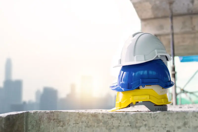 Tres cascos de seguridad apilados (blanco, azul y amarillo) sobre un muro de concreto con la ciudad al fondo