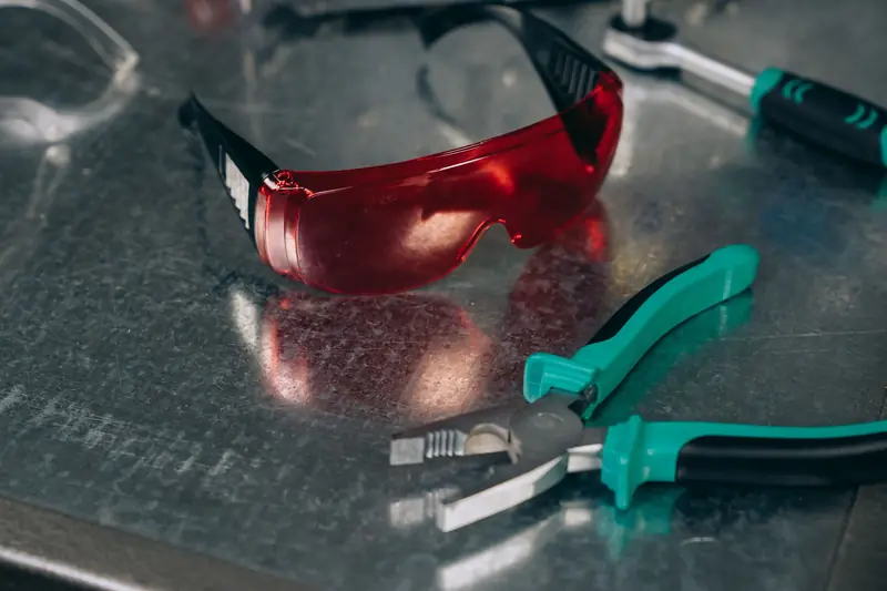 Lentes de seguridad de luna roja junto a un alicate sobre una mesa metálica de taller