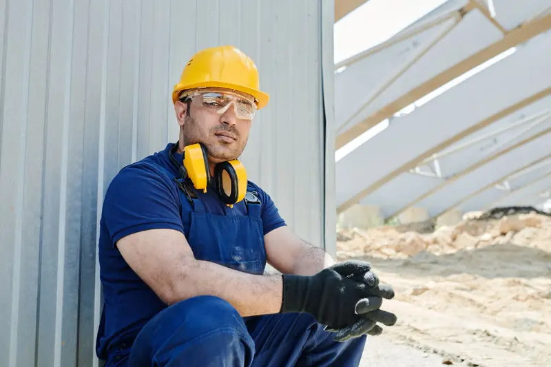 Trabajador con overol azul tipo jardinero, casco amarillo, lentes, orejeras al cuello y guantes, sentado junto a una pared de calamina
