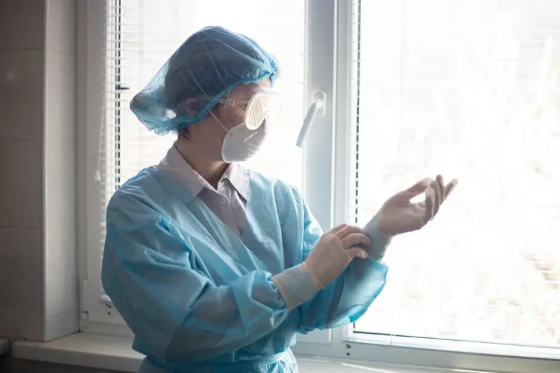 Trabajadora de salud con bata descartable celeste, gorro, respirador descartable y gafas de protección cerradas, ajustándose los guantes de látex sobre los puños de la bata junto a una ventana