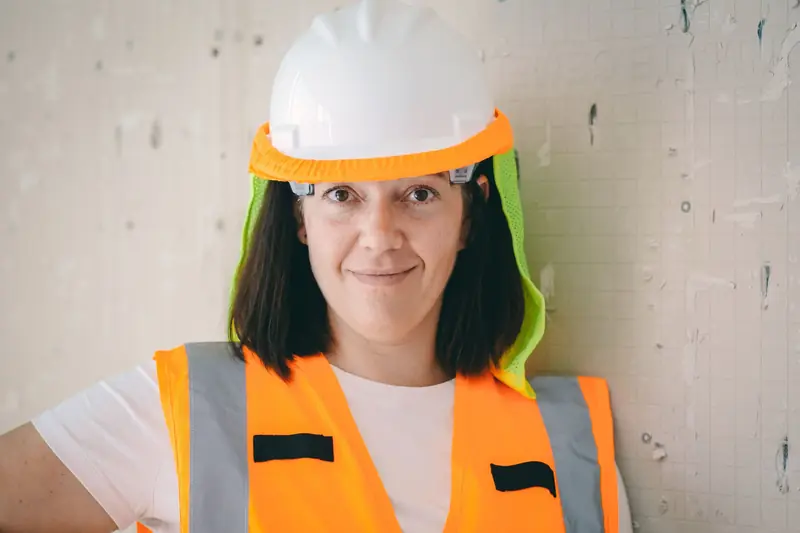 Trabajadora con casco blanco y cubrenuca de malla verde fosforescente sujeto con banda naranja, y chaleco reflectivo naranja, apoyada en una pared de obra