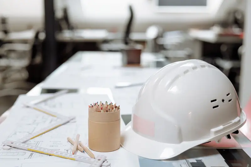 Casco blanco sobre una mesa de oficina con planos, regla plegable y lápices de colores