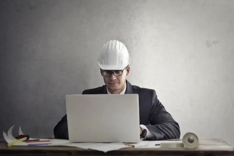 Ingeniero con casco blanco y lentes revisando registros en una laptop sobre una mesa con planos