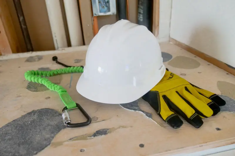 Casco blanco, guantes de trabajo y eslinga con mosquetón dispuestos sobre una mesa para su revisión.