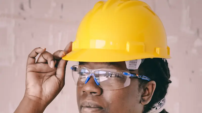 Trabajadora con casco amarillo, lentes de seguridad transparentes y chaleco de alta visibilidad, ajustándose el casco con la mano