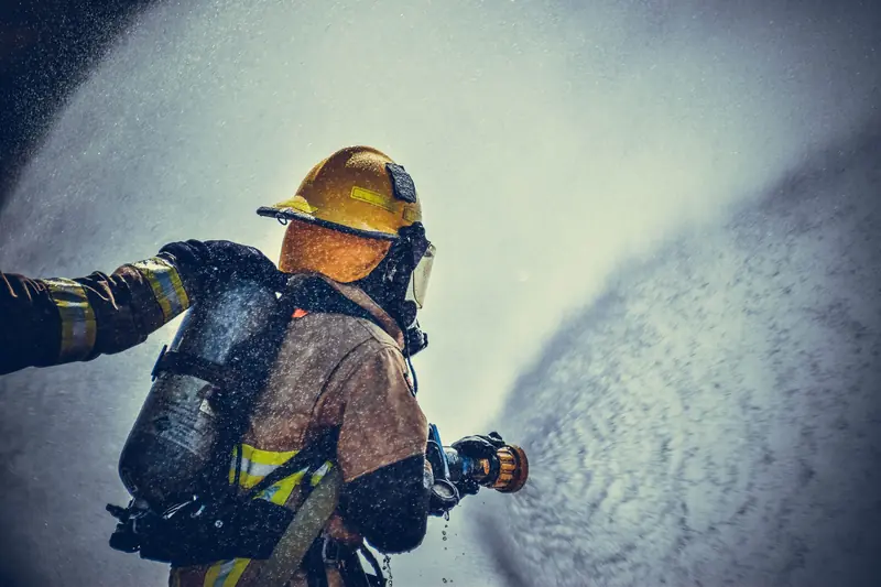 Bombero de espaldas con casco amarillo, cubrenuca naranja, máscara de respiración autónoma, chaquetón con cintas reflectivas, cilindro de aire y guantes, lanzando agua con una pitón; a la izquierda se ve el brazo de otro bombero