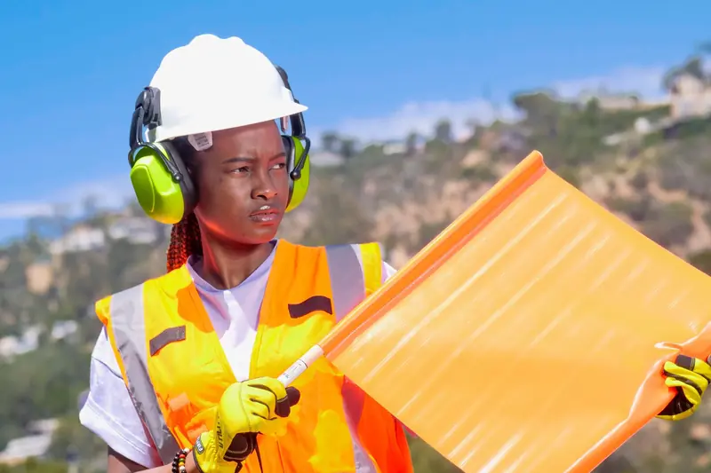 Trabajadora con casco blanco, orejeras, chaleco reflectivo y guantes sosteniendo una bandera de señalización