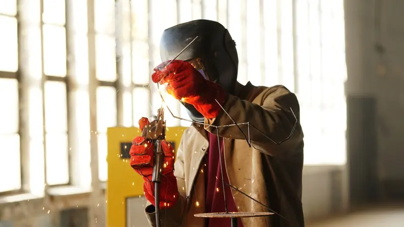 Soldador con careta de soldar, guantes de cuero rojos y casaca de cuero trabajando en un taller iluminado