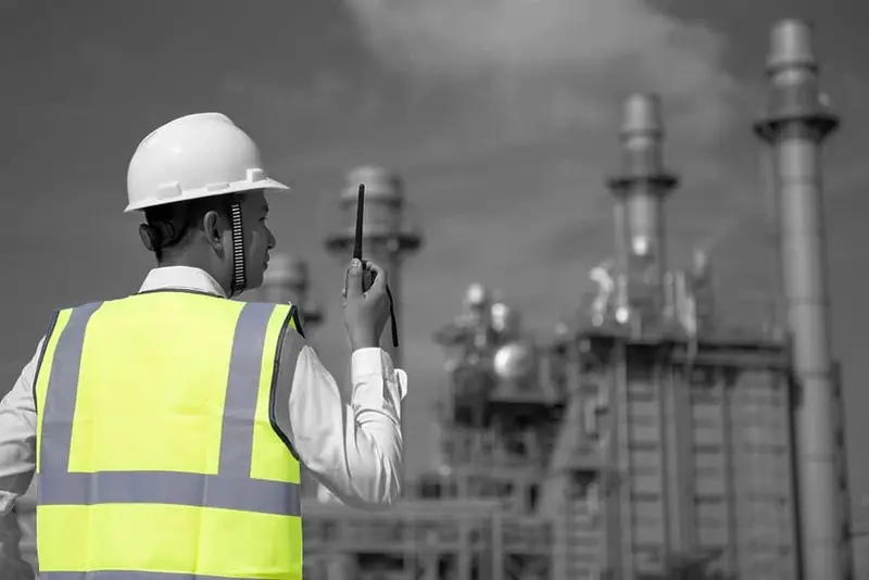 Supervisor de espaldas con casco blanco y chaleco reflectivo amarillo frente a una planta industrial, en blanco y negro con el chaleco en color