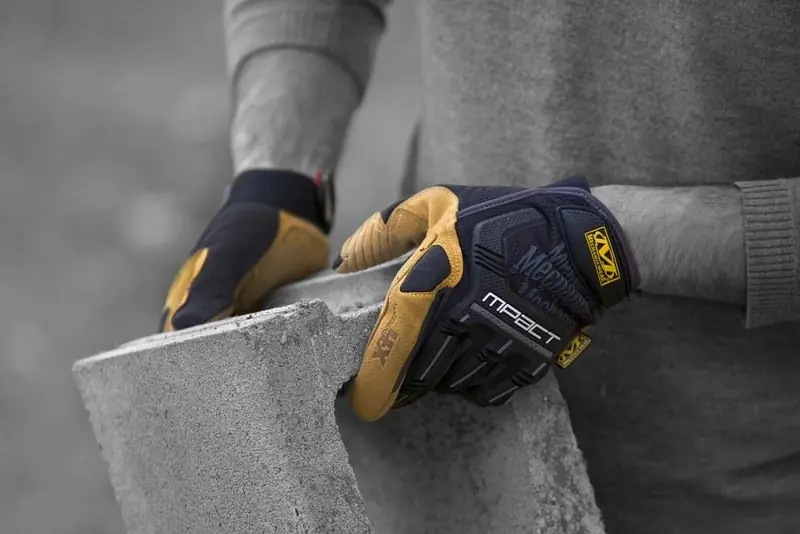 Manos con guantes de trabajo con protección en el dorso sosteniendo un bloque de concreto, en blanco y negro con los guantes en color