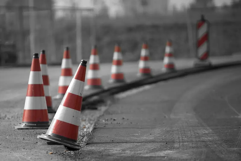 Fila de conos de seguridad naranjas con franjas blancas delimitando el borde de una vía, en blanco y negro con los conos en color