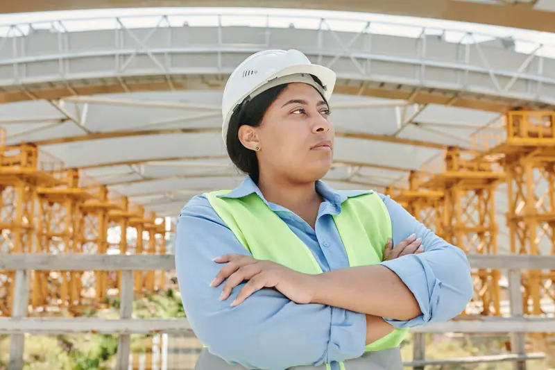 Ingeniera con casco blanco y chaleco de alta visibilidad, de brazos cruzados bajo una estructura metálica en construcción