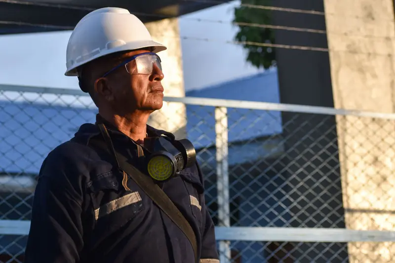 Trabajador con casco blanco, lentes de seguridad y respirador de media cara colgado al cuello, con uniforme con cintas reflectivas, de pie junto a una malla de obra